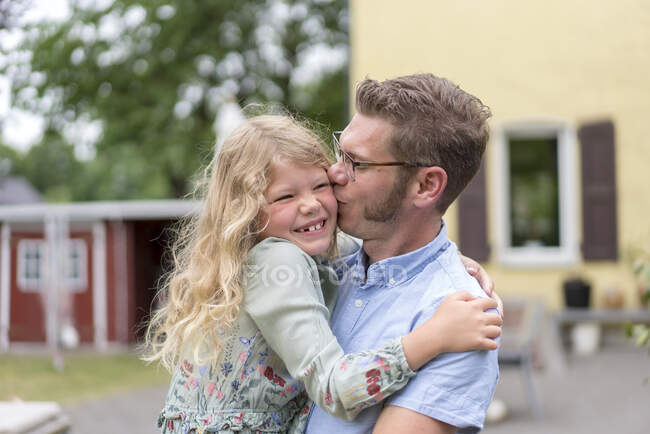 Мужчина целует счастливую блондинку дочь, неся ее на заднем дворе — стоковое фото