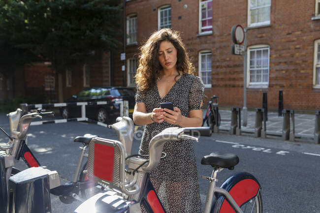 Beautiful woman renting bicycle through smart phone at parking station - foto de stock