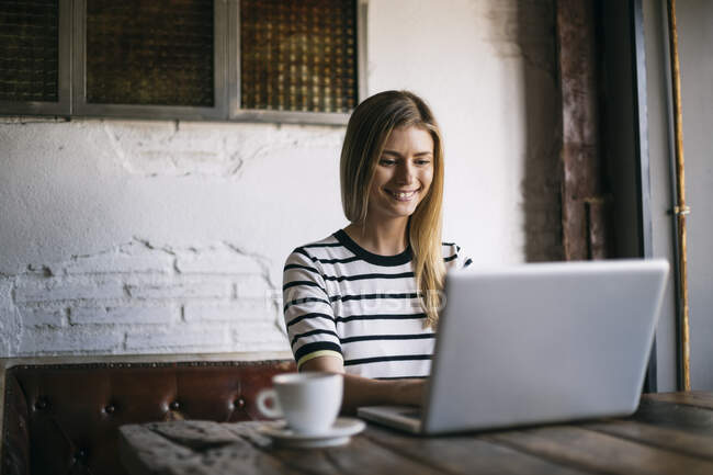Mujer adulta sonriendo mientras usa el portátil en la cafetería - foto de stock