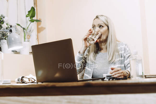 Mujer mayor bebiendo agua durante la consulta en línea en casa - foto de stock