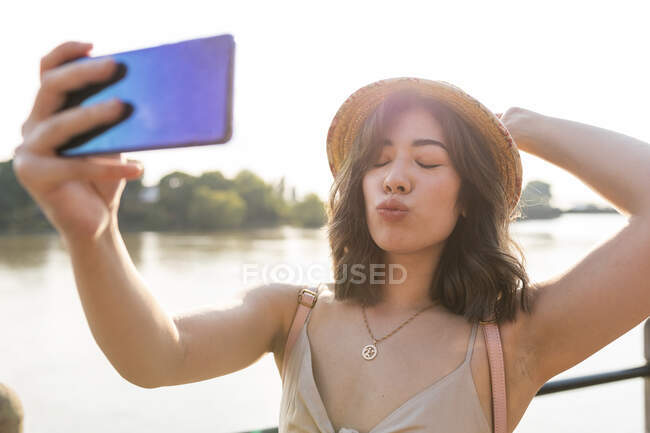 Beautiful woman grimacing while taking selfie on smart phone standing against river — Stock Photo