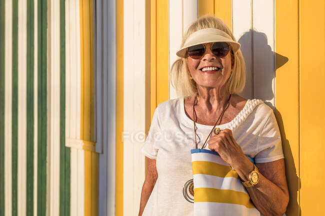 Modische Seniorin mit Augenschutz und Sonnenbrille steht vor bunt gestreifter Wand — Stockfoto