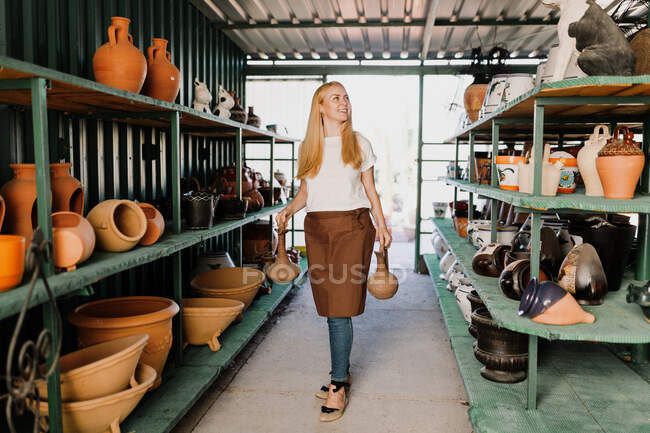 Усміхаючись, жінка тримає кераміку, стоячи на полицях у майстерні. — стокове фото