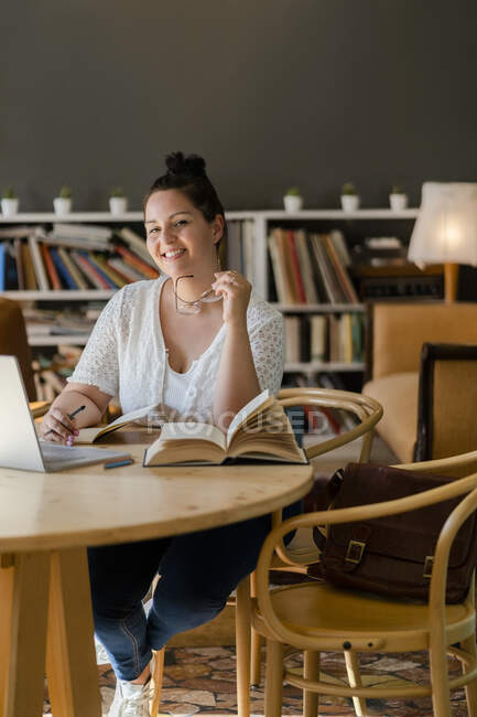 Mujer joven sonriente con libros y portátil en la mesa estudiando en la cafetería - foto de stock