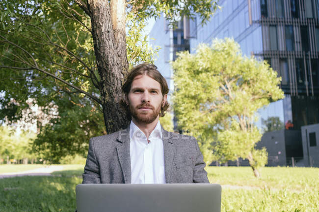 Confident handsome businessman sitting with laptop against tree at financial district - foto de stock
