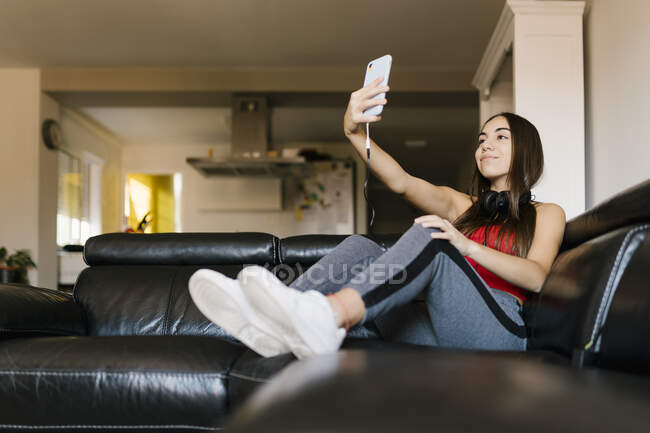 Sorridente ragazza adolescente che prende selfie attraverso il telefono cellulare mentre si siede sul divano a casa — Foto stock