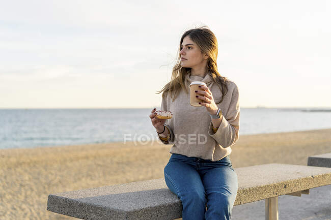 Nachdenkliche junge Frau hält frischen Donut mit Einwegbecher in der Hand, während sie am Strand auf einer Bank sitzt und bei Sonnenuntergang wegschaut — Stockfoto
