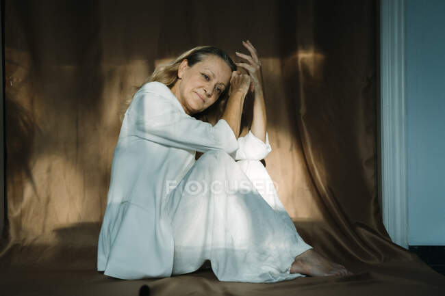 Mujer mayor pensativa sentada contra cortina marrón - foto de stock
