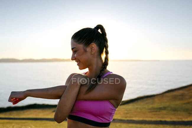 Усмішка молода жінка розтягуючи руки, стоячи проти моря на заході сонця. — стокове фото