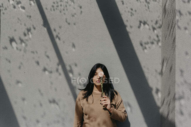Femme tenant fleur devant les yeux tout en se tenant contre un mur de béton par une journée ensoleillée — Photo de stock