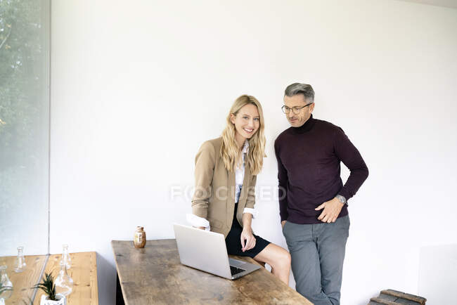 Mujer de negocios sonriente discutiendo sobre portátil con compañero de trabajo masculino contra la pared en la oficina - foto de stock