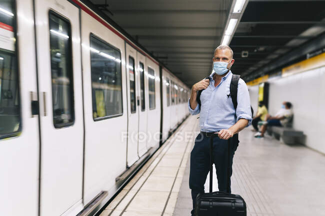 Empresario con máscara facial mientras está de pie con el equipaje en la plataforma del metro - foto de stock
