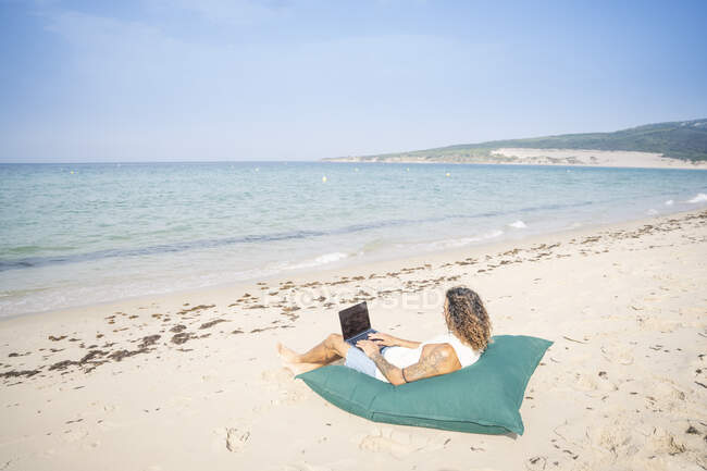 Uomo che utilizza il computer portatile mentre si rilassa su un grande cuscino in spiaggia, Tarifa, Spagna — Foto stock