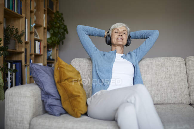 Mujer con las manos detrás de la cabeza escuchando música a través de auriculares en casa - foto de stock