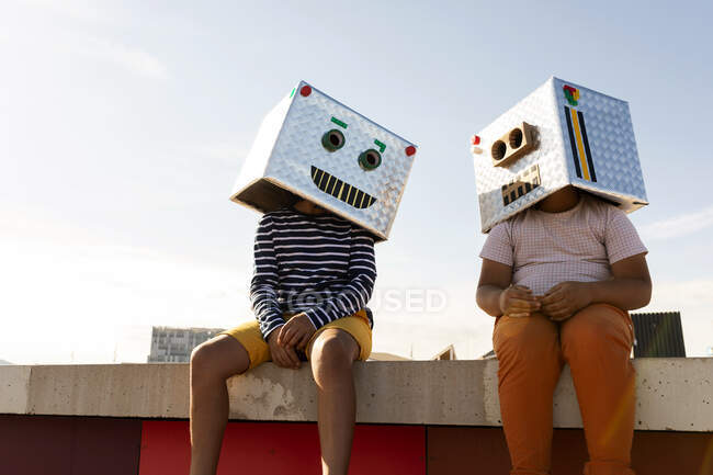Friends wearing robot masks sitting on retaining wall against clear sky during sunny day - foto de stock