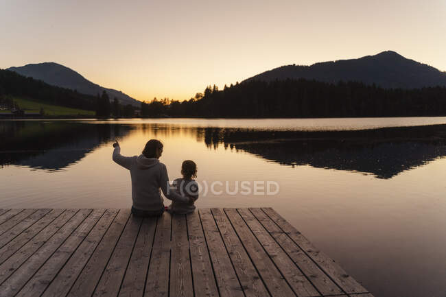 Mother and little daughter sitting together at end of lakeshore jetty at dusk — Stock Photo