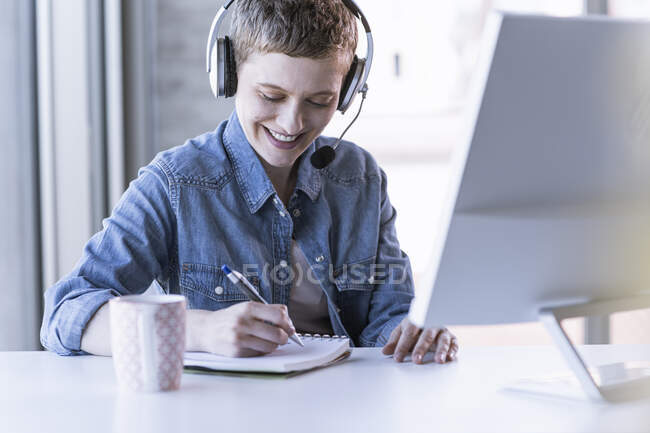 Smiling businesswoman wearing headset at desk in office taking notes - foto de stock