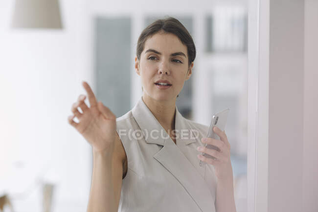 Mujer joven apuntando a la pared de vidrio mientras usa el teléfono móvil en la oficina - foto de stock