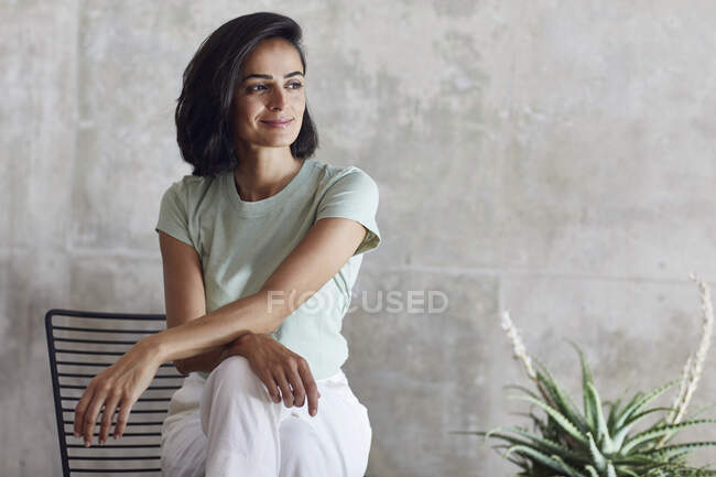 Pensativa mujer de negocios sonriendo mientras se sienta en la silla contra la pared gris en la oficina - foto de stock