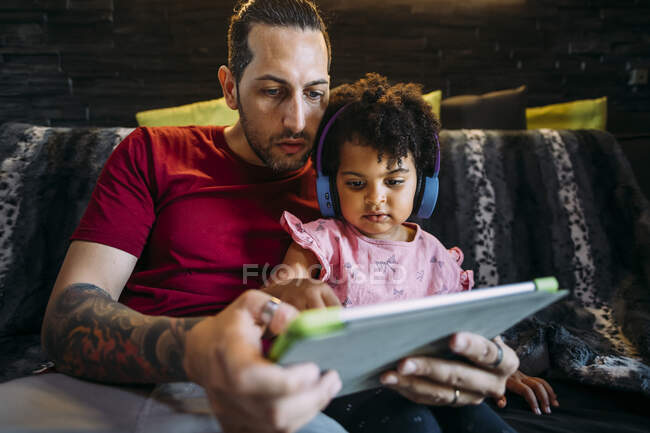 Отец с дочерью используют цифровой планшет, сидя дома на диване — стоковое фото