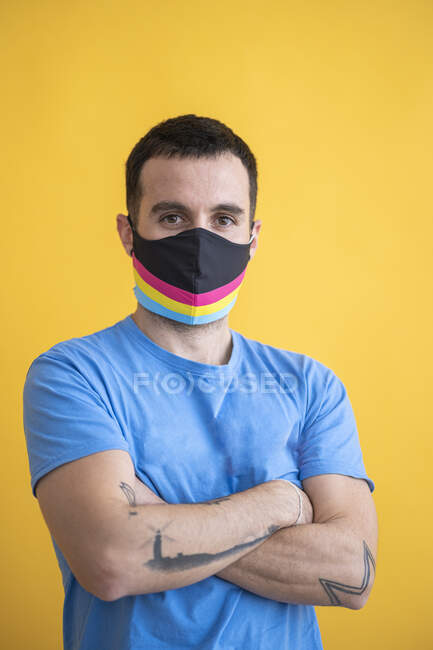 Gros plan de l'homme portant un masque multicolore avec les bras croisés debout sur fond jaune — Photo de stock