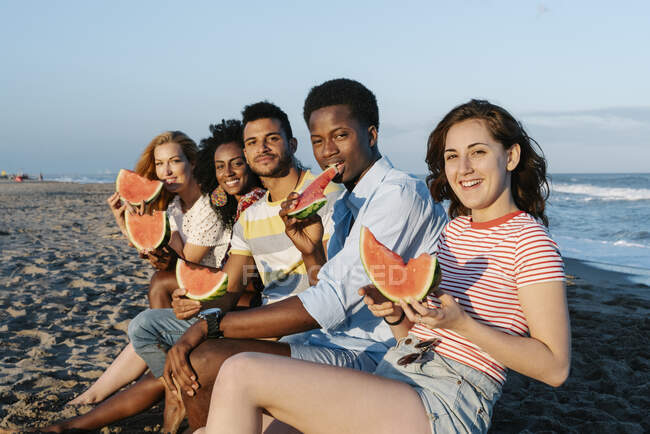 Улыбающиеся друзья едят арбуз, сидя на пляже в солнечный день — стоковое фото