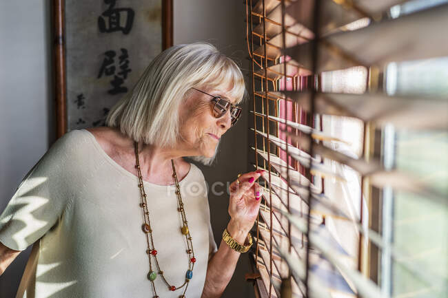 Seniorin mit Sonnenbrille schaut zu Hause durch Fenster — Stockfoto