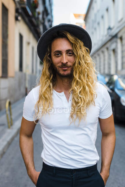 Стильный мужчина средних лет с длинными волосами в шляпе, стоя на улице в городе — стоковое фото