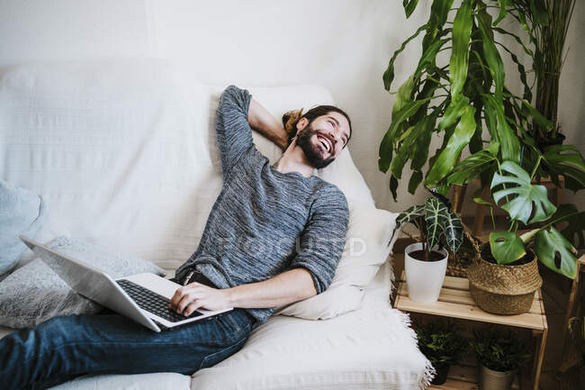 Smiling man with laptop looking away while sitting on sofa at home — Stock Photo