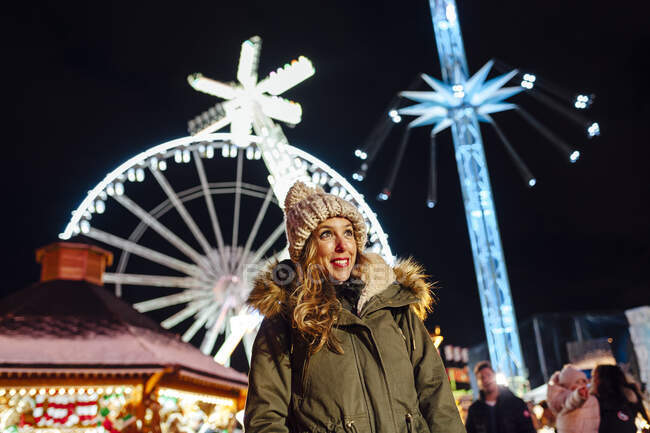Sorrindo mulher olhando para longe, enquanto de pé contra passeios de parque de diversões à noite durante o Natal — Fotografia de Stock