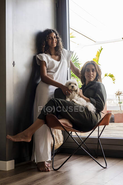 Дівчина, що стоїть біля жінки, тримає цуценя, сидячи на стільці вдома — стокове фото