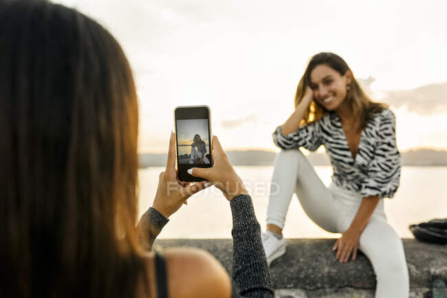 Woman taking photo of friend sitting on retaining wall at promenade — Stock Photo