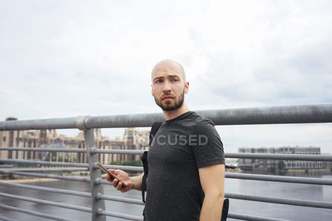 Young man using smart phone looking away while standing by railing against sky — Stock Photo