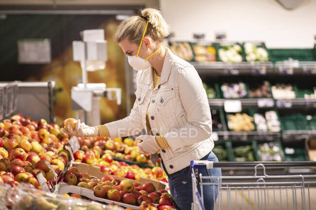 Дівчинка підліткового віку в захисній масці і рукавичках вибирає яблука в супермаркеті. — стокове фото