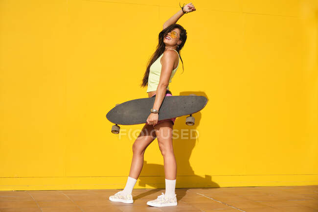 Portrait of young woman standing in front of yellow wall with longboard in hand — Foto stock
