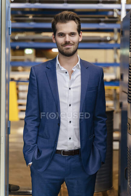Jovem empresário vestindo terno de pé com as mãos nos bolsos na fábrica — Fotografia de Stock