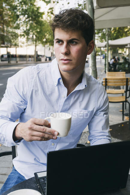 Продуманий молодий чоловік тримає чашку кави, сидячи з ноутбуком у тротуарному кафе — стокове фото