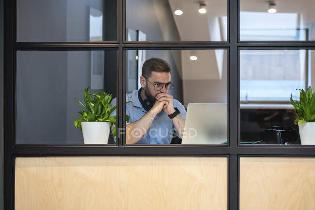 Thoughtful handsome male entrepreneur looking at laptop seen through glass wall in illuminated office — Foto stock