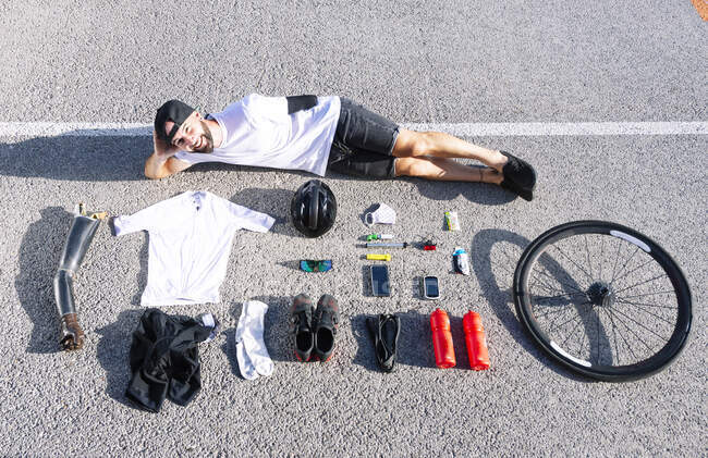 Athlète adaptatif masculin souriant avec divers objets allongés sur la route — Photo de stock