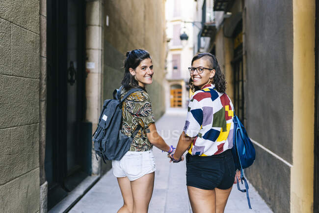 Fidanzate sorridenti che si tengono per mano guardando oltre la spalla mentre camminano sul vicolo in città — Foto stock