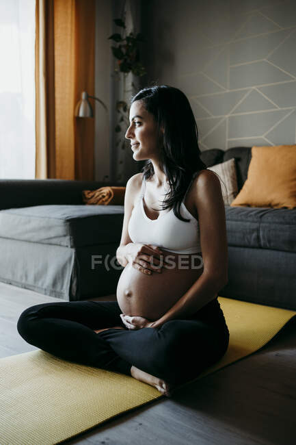 Thoughtful pregnant woman holding stomach while sitting on exercise mat at home — Stock Photo