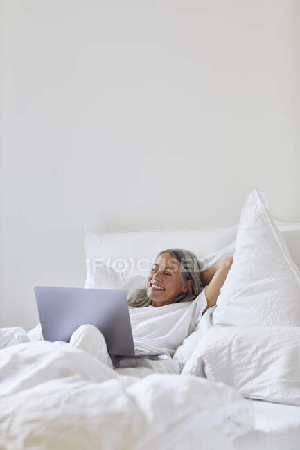 Mujer sonriente usando el ordenador portátil mientras está acostado en la cama en casa - foto de stock
