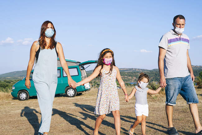 Familia con máscaras cogidas de la mano mientras camina sobre el paisaje contra el cielo - foto de stock