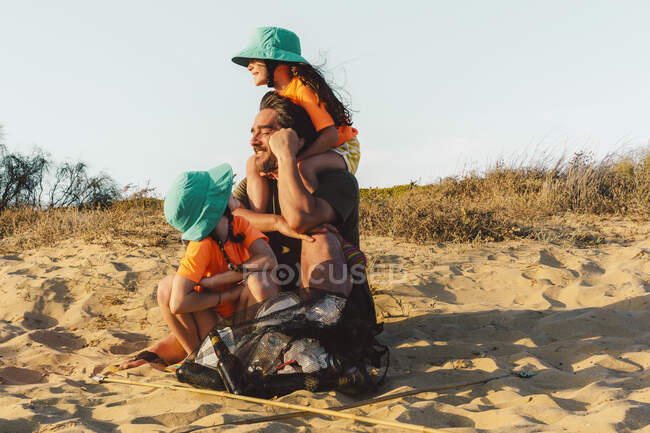 Padre e hijas sentados en la arena con bolsa de basura durante el día soleado - foto de stock