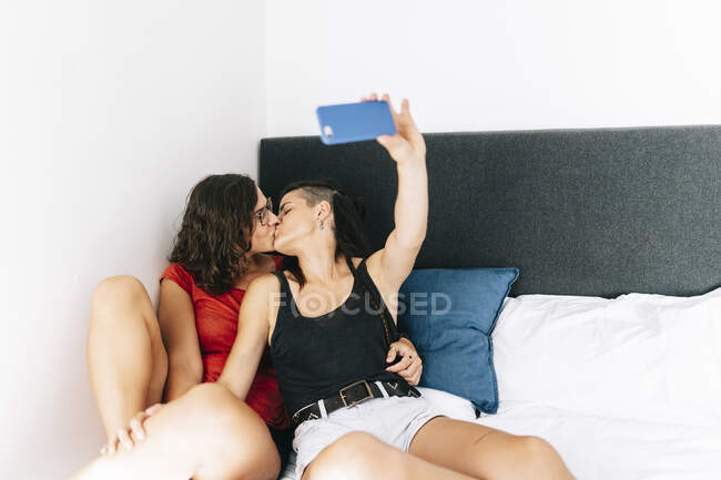 Молодые подруги целуются, делая селфи на смартфоне дома — стоковое фото