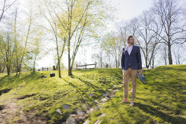 Thoughtful male professional looking away while standing barefoot on grass against trees at park — Stock Photo