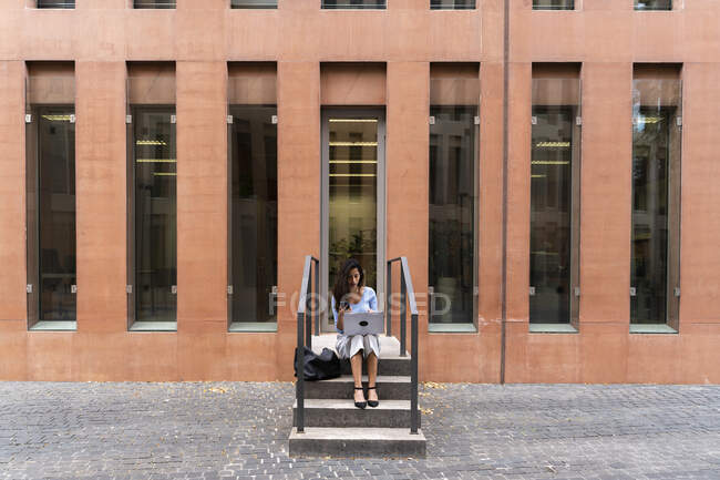 Зайнята жінка користується ноутбуком, сидячи на сходах при вході до офісного будинку. — стокове фото