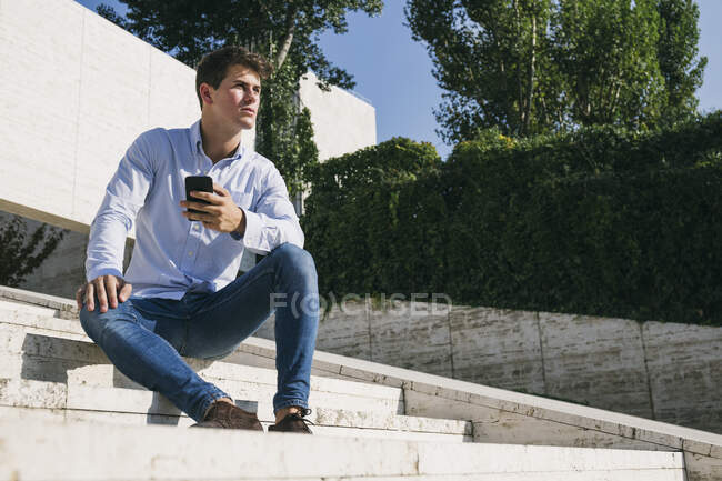 Beau jeune homme réfléchi assis avec un téléphone intelligent sur les marches contre les arbres en ville pendant la journée ensoleillée — Photo de stock