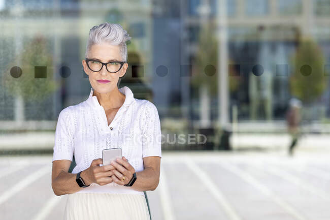 Старша жінка, яка користується розумним телефоном, стоячи проти скла на вулиці. — стокове фото