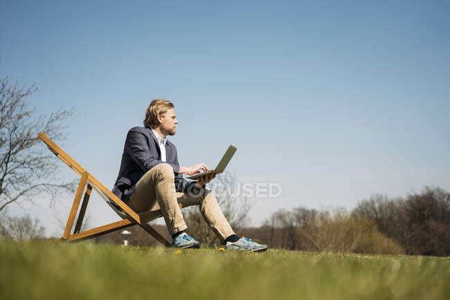 Superficie vista livello di uomo d'affari guardando lontano mentre seduto con computer portatile sulla sedia a parco durante la soleggiata giornata estiva — Foto stock